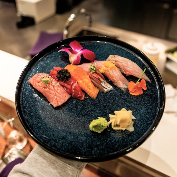 A hand holding a dark blue plate of various Shiso Noodle and Sushi Bar nigiri, garnished with flowers.