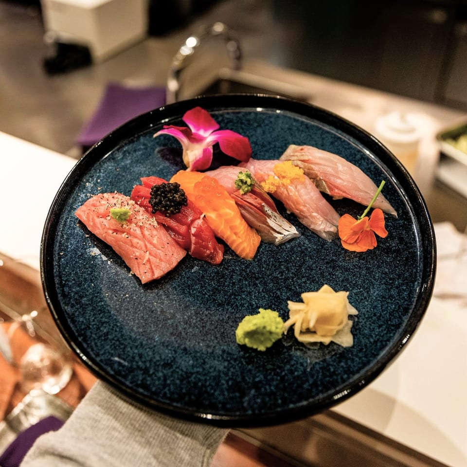 A hand holding a dark blue plate of various Shiso Noodle and Sushi Bar nigiri, garnished with flowers.