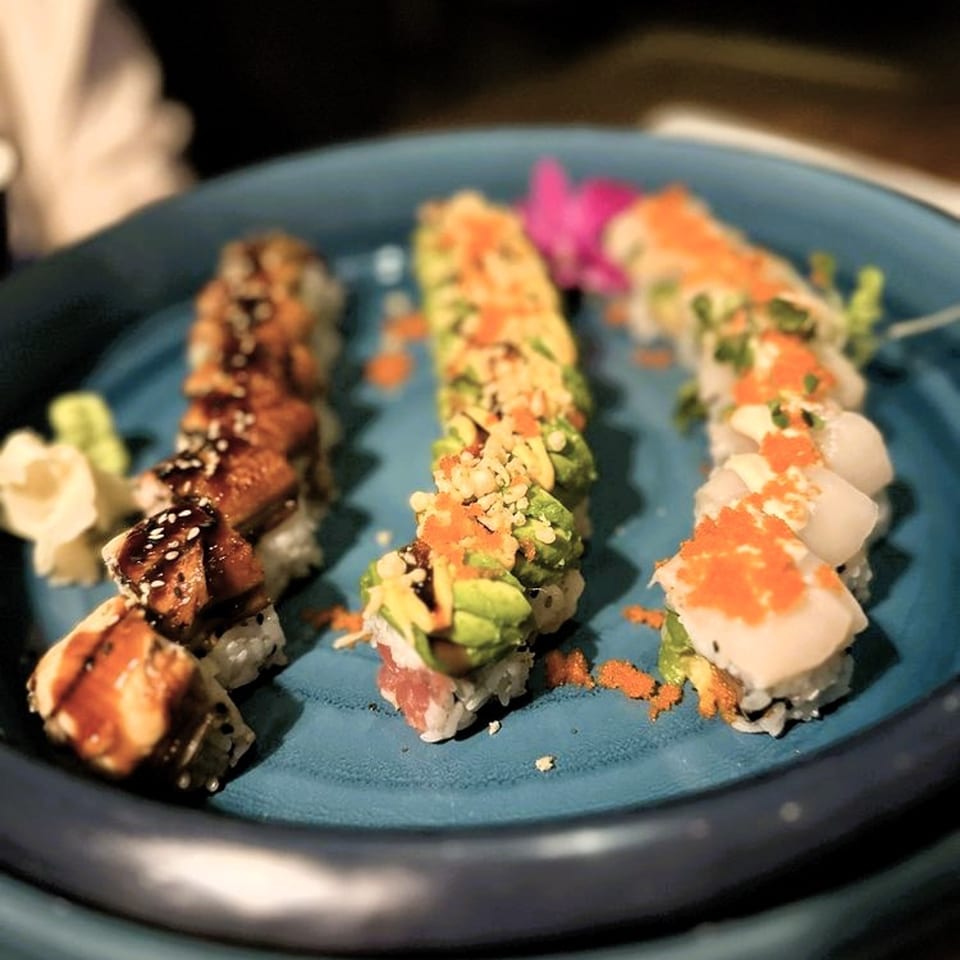 Three rows of sushi rolls with various toppings, including eel, avocado, and roe, on a blue plate.